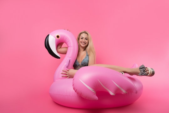 Image of blonde woman in bathing suit and in pink glasses sitting on inflatable flamingo on empty pink background