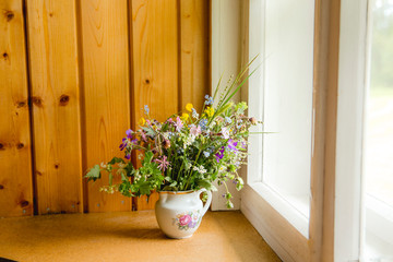 Vintage style flower bouquet made of wild flowers found in forest and meadow, standing in old cream jug on window sill, summers in grandma`s house concept. Northern Europe.