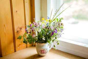 Vintage style flower bouquet made of wild flowers found in forest and meadow, standing in old cream jug on window sill, summers in grandma`s house concept. Northern Europe.