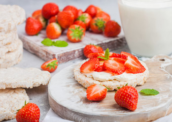 Healthy organic rice cakes with ricotta and fresh strawberries on wooden board and glass of milk on light stone kitchen background. Top view.