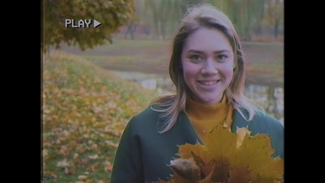 Young woman walks in the autumn park. Cheerful woman with blond hair in a green coat walks on the yellow leaves. Girl with a bouquet of leaves in her hands. Old video. Home video archive. Family video