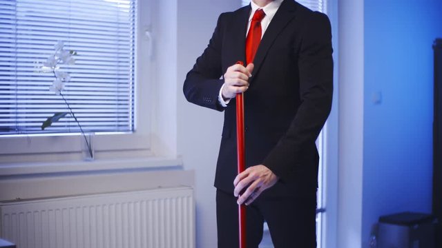 Businessman brush the floor doing housework 4K. Medium shot of headless male businessman in focus while holding a red broom handle. Windows in background.