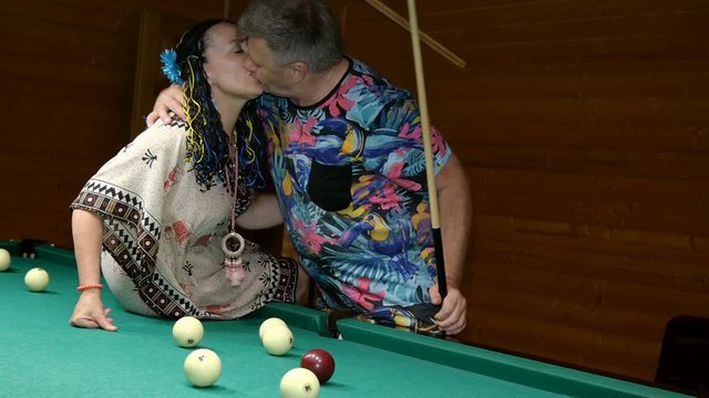 Elderly family. Senior man and woman relax and have fun in billiard room. Huggings, kisses, rejoice and beat balls with cue. Selective focus.