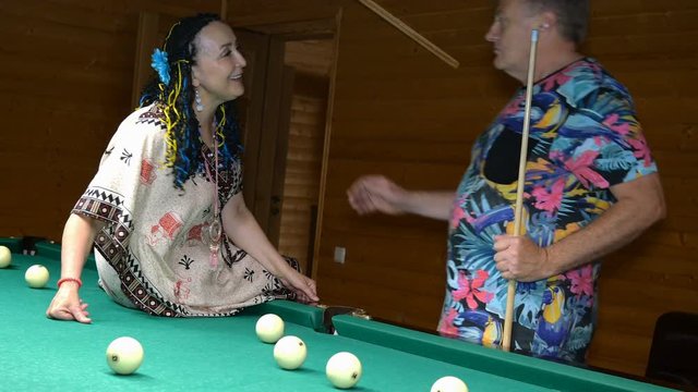 Elderly family. Senior man and woman relax and have fun in billiard room. Huggings, kisses, rejoice and beat balls with cue. Selective focus.