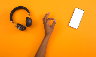 Black girl showing ok gesture, wireless headphones and blank smartphone