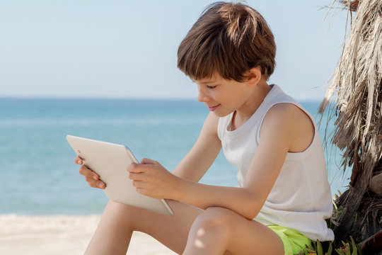 Child Looks At Tablet In The Shade Of A Palm Tree On The Beach