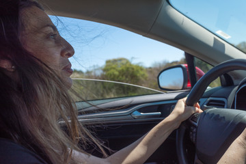 a woman smirks as she drives while the wind blows around her hair with the window down
