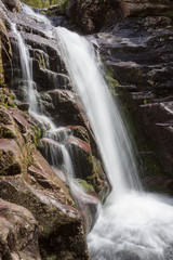 Fototapeta premium Close up view of a scenic waterfall cascading down the red, wet rocky cliff covered by sunlit moss