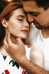 Close up portrait of a amazing sensual couple face to face with closed eyes while man is touching face of her girlfriend with red hair and freckles outside .