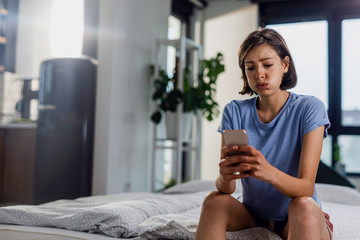 Young displeased woman reading text message on cell phone in the bedroom.