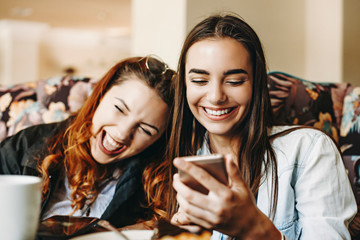 Beautiful young female holding a smartphone laughing while showing something to her girlfriends which is laughing leaning head on her shoulder.