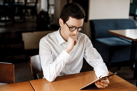 Cute young caucasian man sitting at a desk and looking at a tablet while wearing white shirt and eyeglasses.