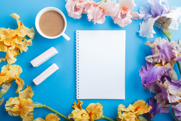 Purple, pink, yellow iris flowers and a cup of coffee with notebook on pastel blue background.