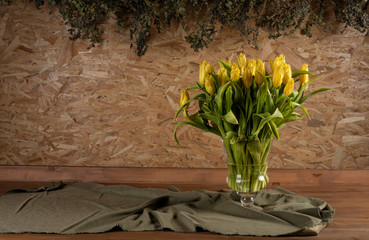 Yellow tulips in the vase on wooden table. Leaves dangling from the OSB board in the background.