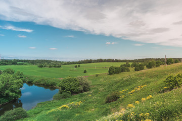 Colorful Bright Sunny Green Field Landscape With Blue Cloudy Sky, River, Trees And Hills
