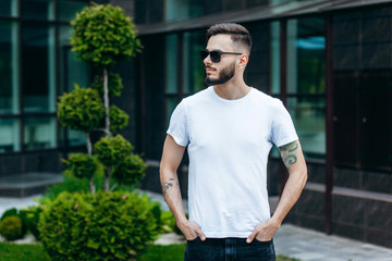 Fototapeta premium A young stylish man with a beard in a white T-shirt and glasses. Street photo