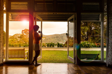 Woman admiring natural scenic sunrise from home interior with coffee