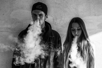 Vape teenager. Young cute girl in casual clothes and handsome guy in a cap smoke an electronic...