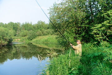 Obraz na płótnie Canvas Fisherman male catch fish on river coast in grass, man in green clothes and hat throws fishing-rod in water, view from side, horizontal stock photo image