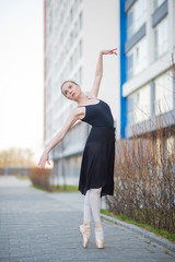 Ballerina in a tutu posing against the backdrop of a residential building. Beautiful young woman in black dress and pointe shoes dancing ballet outside. Elegant classical dance