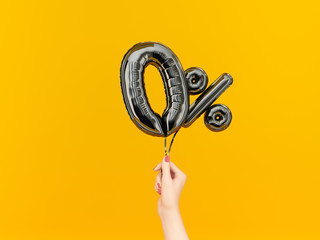 Female hand holding Zero percent symbol. 0 % offer black flying foil balloons on yellow. Loan rate...