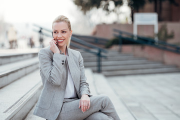 Positive businesswoman in formal wear sitting on the stairs in the city and talking on the phone.