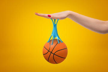 Orange basketball ball stuck to female hand with blue sticky slime on yellow background