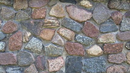 A textured fragment of the relief facade of a wall made of grainy granite stone of brown, gray, beige and maroon hues, fastened with gray cement