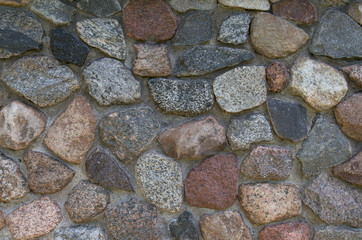 A textured fragment of the relief facade of a wall made of grainy granite stone of brown, gray, beige and maroon hues, fastened with gray cement