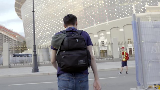 Young man with black backpack walking in city. Action. Rear view of confident guy crossing road with black backpack going to stadium in city