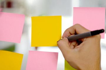 Hand holding pan writing on blank colorful sticky note papers on glass board at office, business stationery supplies