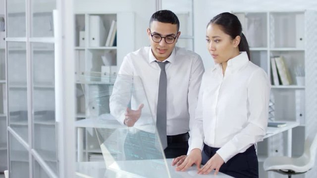 Medium shot of Arab businessman and Asian businesswoman standing together at desk at transparent AR computer display and using it