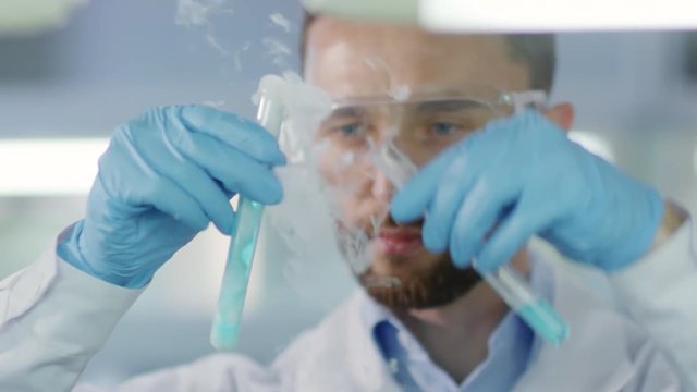 Cinemagraph of male Caucasian scientist in medical uniform, gloves and goggles holding test tube with blue liquid, then pouring it into another test tube and analyzing chemical reaction