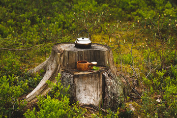 Wooden kuksa with coffee and kettle on the stump in focus. The forest background is blurred