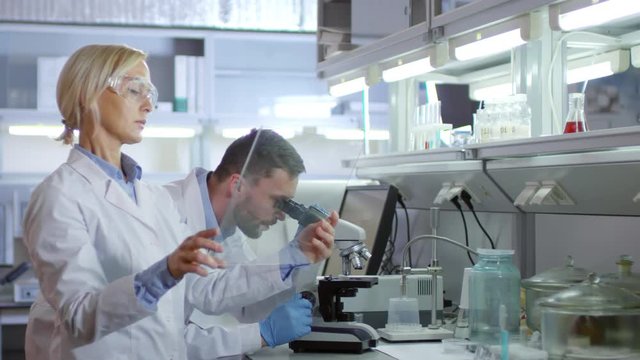 Medium shot of male chemist doing research with microscope and discussing it with his female colleague standing nearby and using gadget with transparent AR screen
