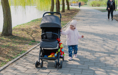 A little girl is rolling baby carriage in the park. Child in the park playing with pram.