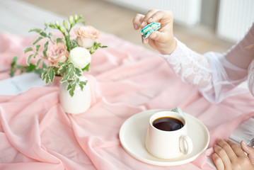 Fototapeta na wymiar Young beautiful caucasian bride enjoying breakfast from french macaroon and coffee on a wooden table with a chiffon pink tablecloth and a vase of flowers.