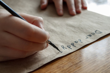 A small girl writes a letter to dear daddy by black paint on brown paper. Closeup, selective focus