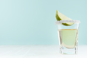 Tequila cocktail with fresh piece lime, rim of salt in shot glass on white wood table and blue...