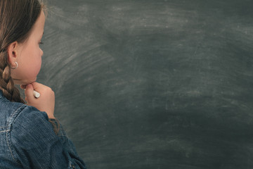 School and education. Back view of thoughtful young girl standing with white chalk at blank...