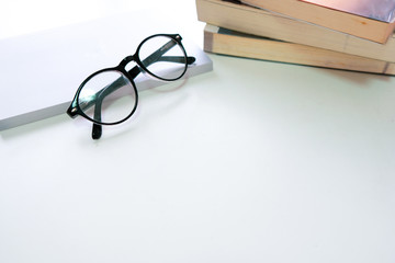 Black eye glasses are placed on a stack of books on a white wooden table beside the window.