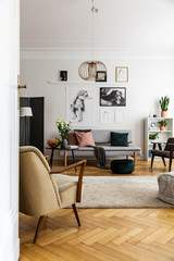Armchair next to carpet and flowers in living room interior with posters above sofa. Real photo
