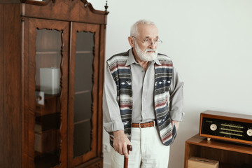 Sad senior man with walking stick standing alone at home