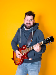 man with black hair and beard, wearing grey hoodie playing the electric guitar in front of a yellow background