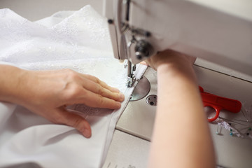 Clothier woman stitching white dress on sewing machine at workplace with pins, threads, scissors on background. Seamstress hands holding and sewing zipper on textile. Blurred background. Close up view