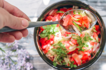 the female hand holds a metal spoon with vegetable salad from tomato, cucumbers and a basil against the background of a clay cup with salad and a wooden old table