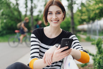 junge frau sitzt auf einer bank im park und hält ihr mobiltelefon in der hand