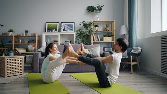 Happy young people exercising at home doing yoga for couples sitting on floor holding hands and putting feet together. Family and healthy lifestyle concept.