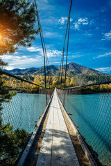 Pedestrian suspended wooden bridge over mountain river. Long bridge across the river against background of mountains covered with forest, golden autumn, indian summer warm sunny day.