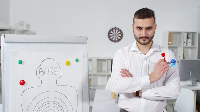 Dollying portrait shot of 20-something company employee, dressed in white shirt, posing next to flipchart with boss target poster, with darts in his hand, staring straight at camera